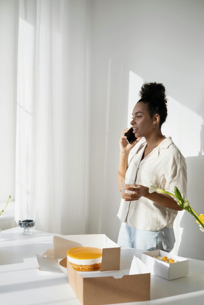African American businesswoman talking on phone with cake packages during work in bright office.